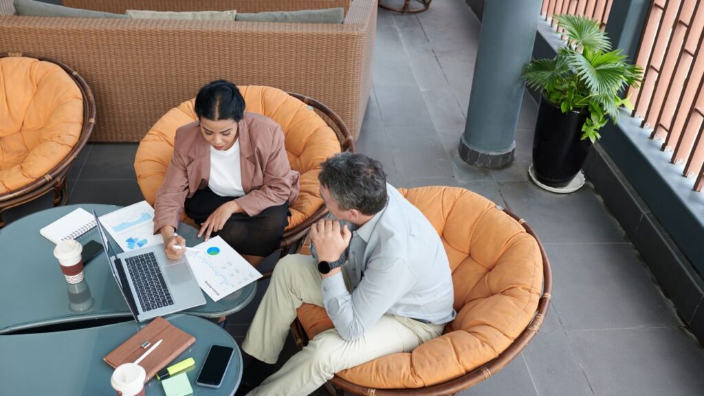 Obrazek przedstawia rozmawiających ze sobą dwoje ludzi, którzy siedzą przy laptopie i popijają kawę