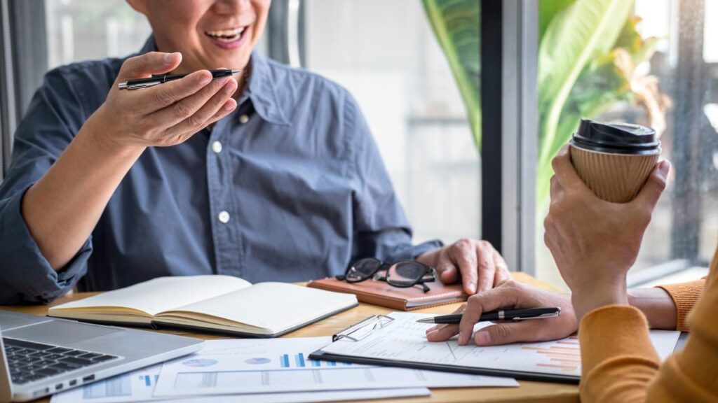 Obrazek przedstawia dwoje rozmawiających ze sobą ludzi, którzy są zadowoleni. Przed sobą mają mnóstwo dokumentów z różnymi projektami i kawę