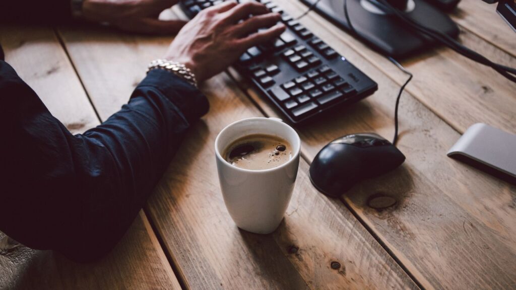 Obrazek przedstawia pracującą przy laptopie kobietę, która popija kawę